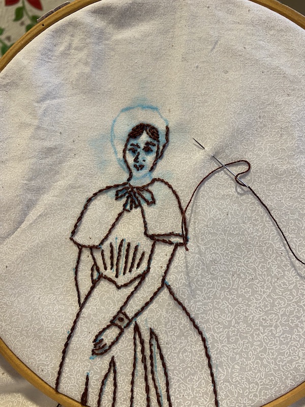 Fabric with hand embroidered figure of a woman in quaker dress from the 1800s. Some of the figure is drawn in blue water erase marker and a needle and thread are stuck in the cloth.