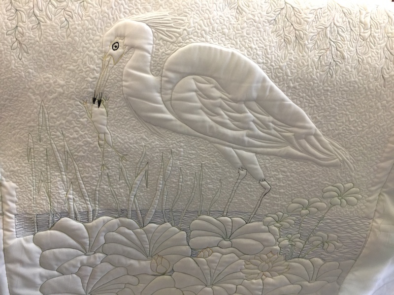 Stitched image of an egret catching a frog. Black outlines on a white fabric.