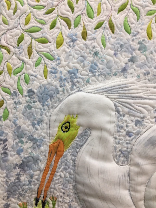 Art quilt detail photo showing the head of an egret with crown of feathers. The background is filled with leaves and mottled grey texture.