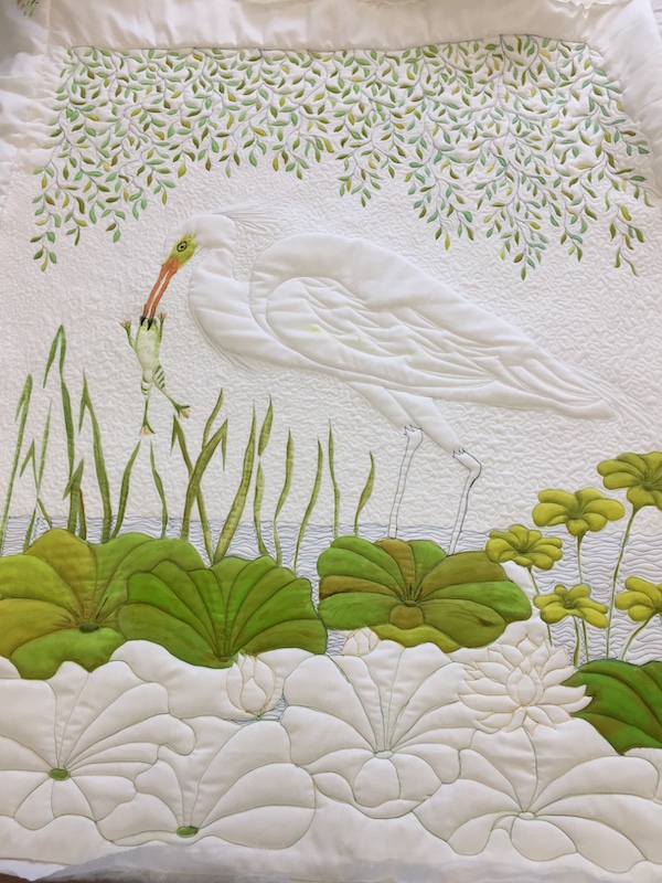Stitched and painted art quilt in progress showing egret standing in lily pads. The lily pad and leaves hanging from the trees above are painted green and large areas are still unpainted.