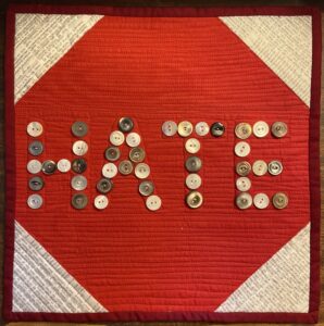 Mini art quilt with red stop sign shape and the letters HATE spelled out in buttons