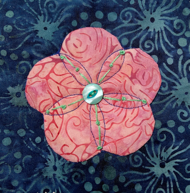 Pink flower with hand embroidery appliquéd onto a dark blue background with a button in the center.