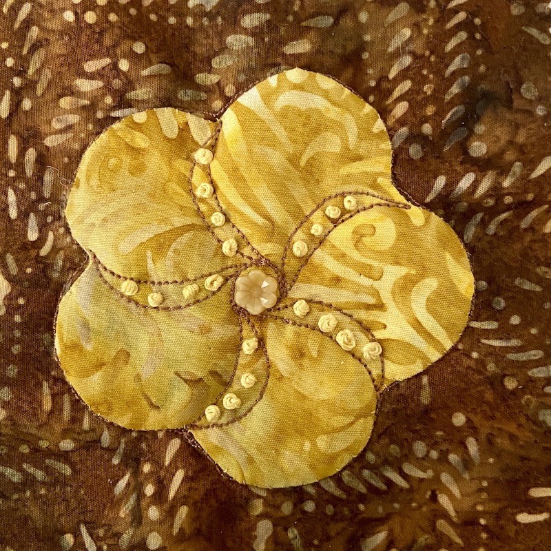 Yellow flower with hand embroidery appliquéd onto a brown background with a button in the center.