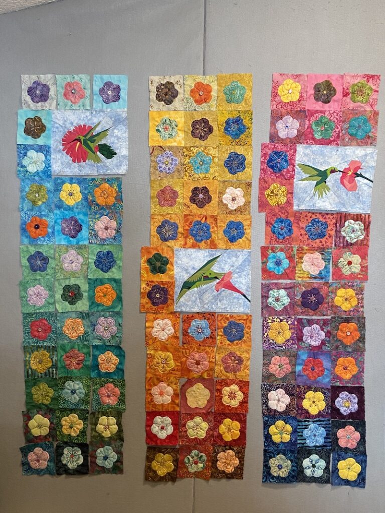 Hummingbird and flower quilt blocks arranged in three long panels with blue on the left, yellow in the middle and pink on the right.