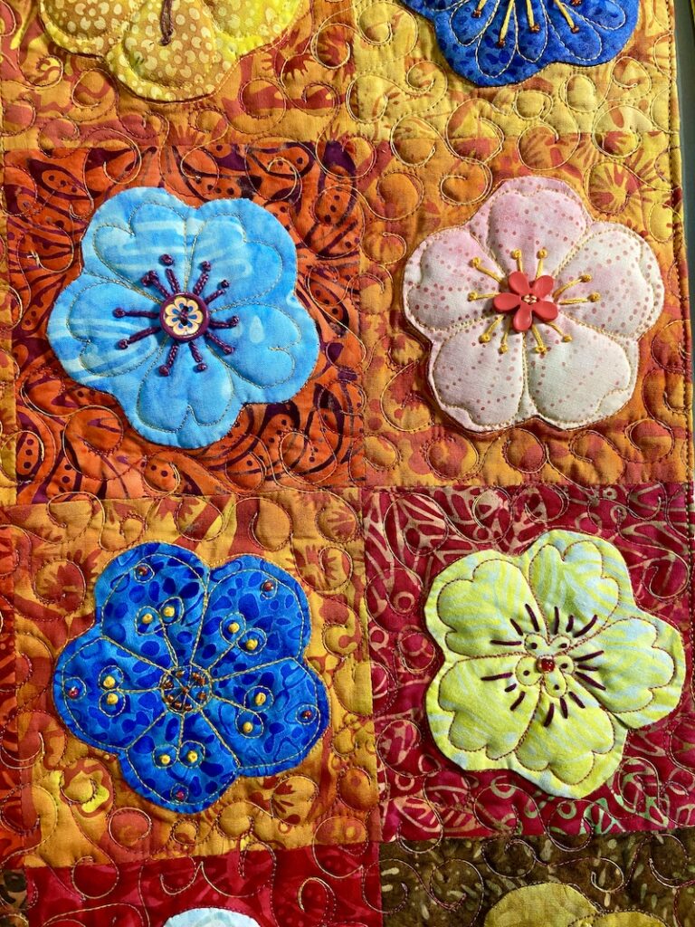 Four appliquéd flowers on orange backgrounds with hand embroidery on the flowers and machine quilting in the background.