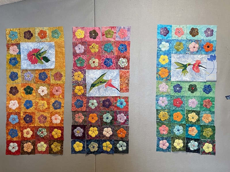 Hummingbird and flower quilt blocks arranged in three shorter panels with yellow on the left, pink in the middle and blue on the right.